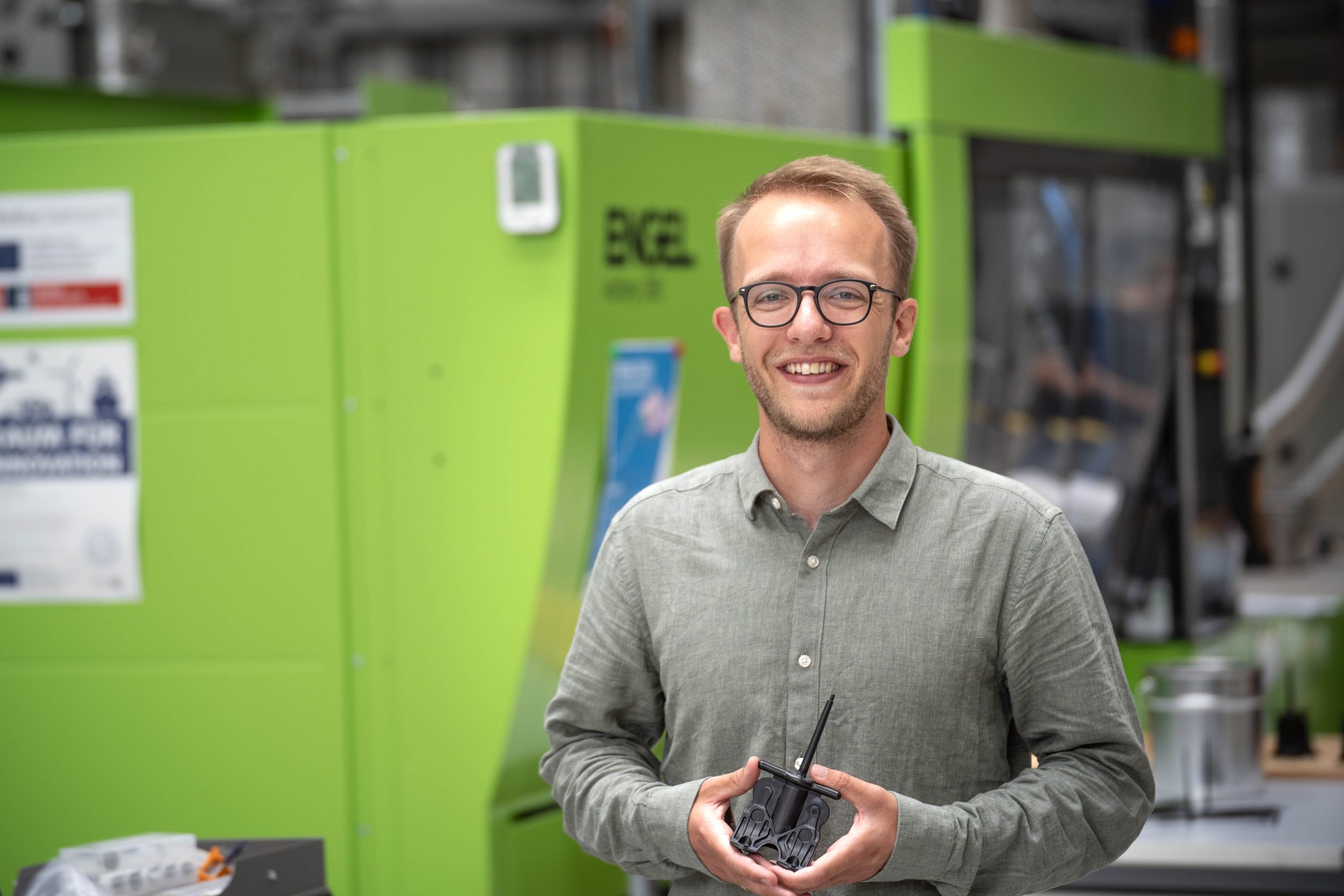 Ein Mann steht vor einer grünen Maschine und hält ein Produkt aus Kunststoff in der Hand
